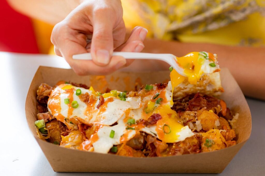 A forkful of runny egg lifted from tater tots topped with fried eggs, bacon, green onions and sriracha