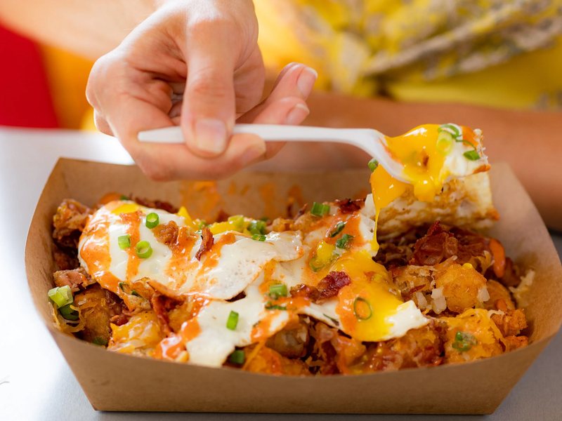 Tater tots topped with fried eggs, bacon, green onions and sriracha, with a runny yolk on the fork